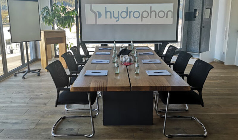 Ein hydrophon Besprechungsraum an dem der Tisch mit Blöcken, Stiften und Getränken gedeckt ist. Im Hintergrund hängt eine Leinwand auf der das Logo von hydrophon gebeamt wird.