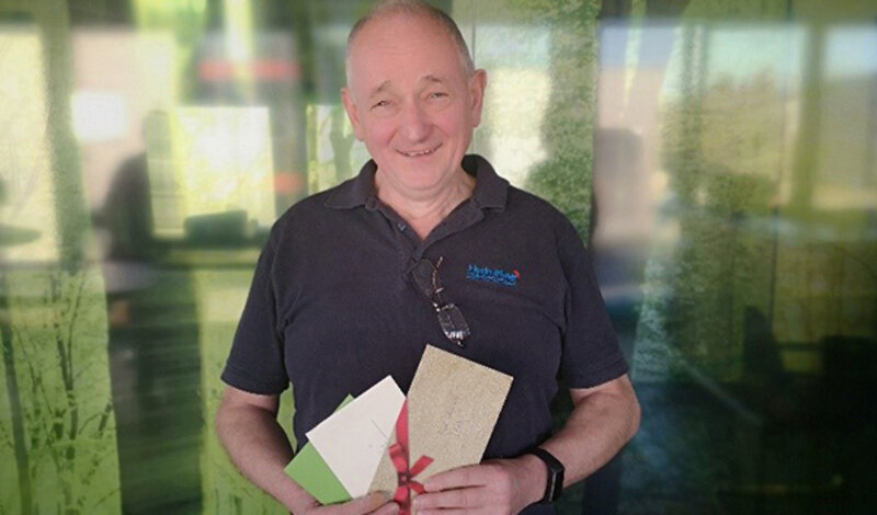 An older man in a hydrophone T-shirt holds envelopes up to the camera and looks at it with a smile.